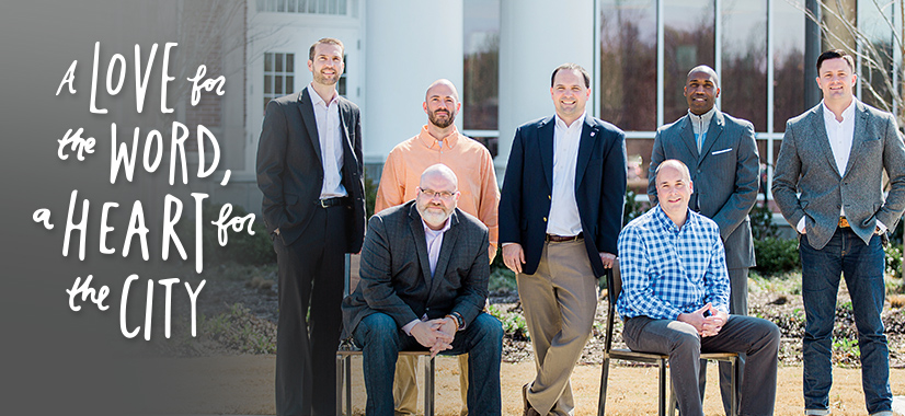 Seven men pose outside a building in business and casual attire. Six are standing and one is sitting. Text on the left reads, “A Love for the Word, a Heart for the City.” Trees and windows are visible in the background.