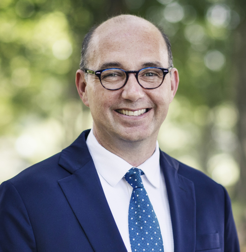 A man wearing glasses, a dark suit, white shirt, and blue patterned tie smiles at the camera in an outdoor setting with blurred greenery at the Union Pennington campus during the Ryan Center Bible Conference.