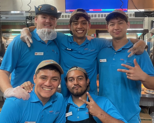 Five restaurant employees in blue uniforms and name tags stand close together, smiling at the camera. Three flash peace signs with their hands. Their camaraderie suggests the teamwork found during a YoungLife summer internship or at Dollywood.