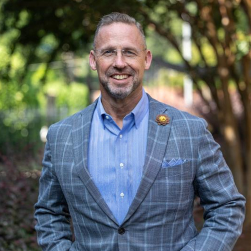 A man with short hair, glasses, and a beard is standing outdoors in a blue shirt and gray plaid suit jacket. He is smiling, with trees blurred behind him—SBC President Pressley of Union Chapel enjoying the greenery.