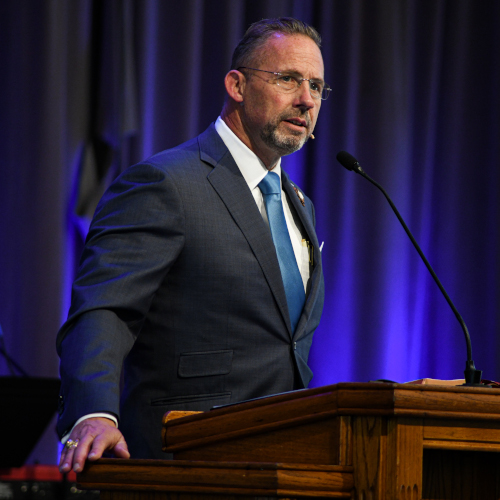 A man in a suit and tie stands at a wooden podium, speaking into a headset microphone. He has short hair, glasses, and a trimmed beard. The background features draped curtains and purple lighting as the SBC President addresses Union students.