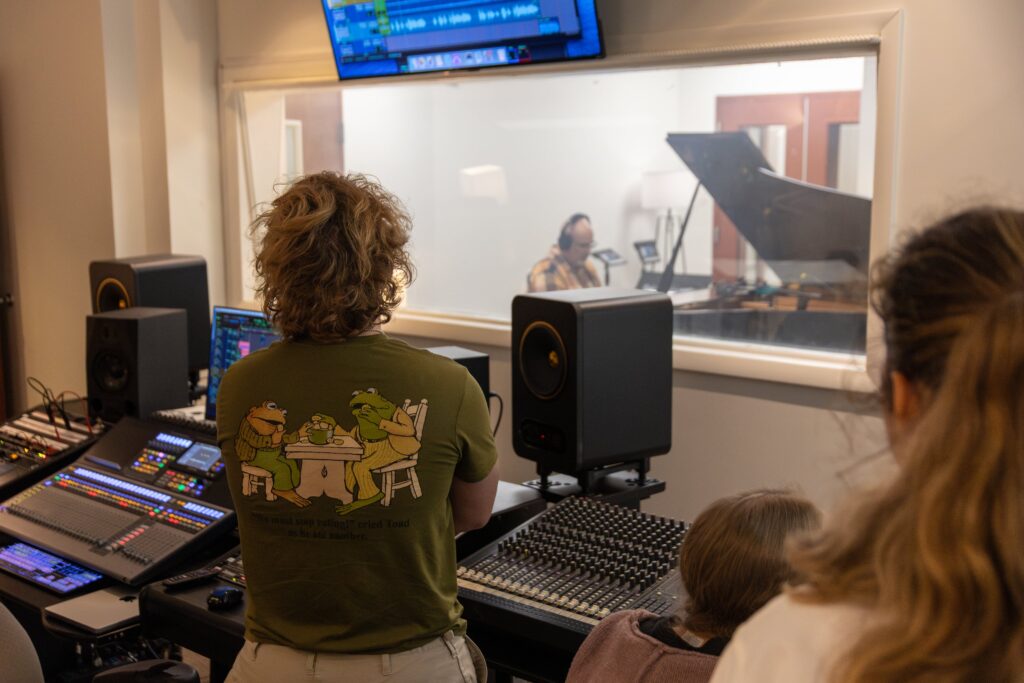 Several people are in a recording studio, with one person at a large 00-Component mixing console and another in front of a piano behind a glass window. Monitors and audio equipment are visible throughout the room.