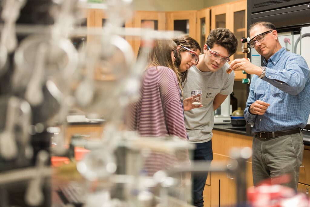 A teacher and two students wearing safety goggles observe a small 00-Component in a science lab, surrounded by lab equipment and wooden cabinets.