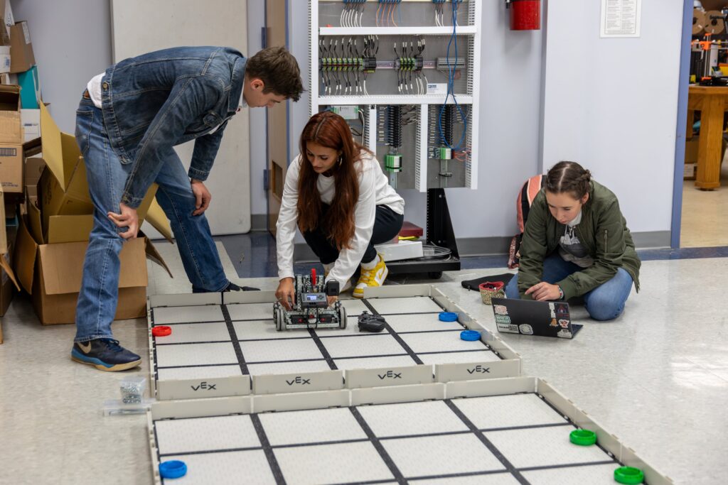 Three people work on a robotics project in a lab. One adjusts a 00-Component robot on a grid mat, another crouches nearby, and the third sits on the floor using a laptop. Shelves with electrical equipment are in the background.
