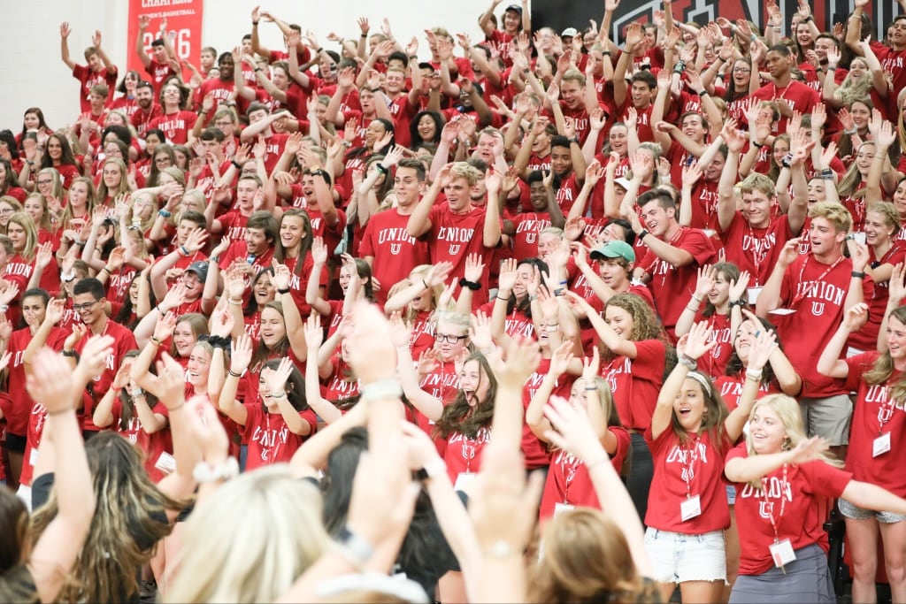 A large component of students wearing matching red Union shirts sit and stand on bleachers, raising their hands and cheering in a crowded indoor gymnasium.