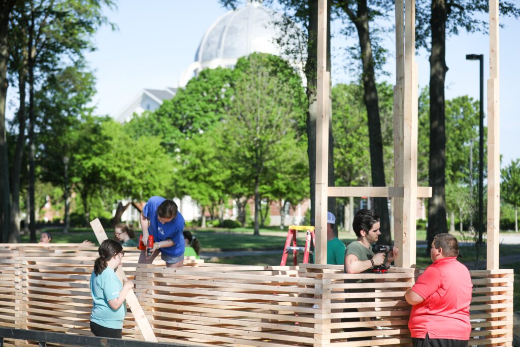 Four people are building a wooden 00-Component structure outdoors in a park-like setting, with trees and a domed building visible in the background. One person uses a power tool while others hold or position wooden slats.