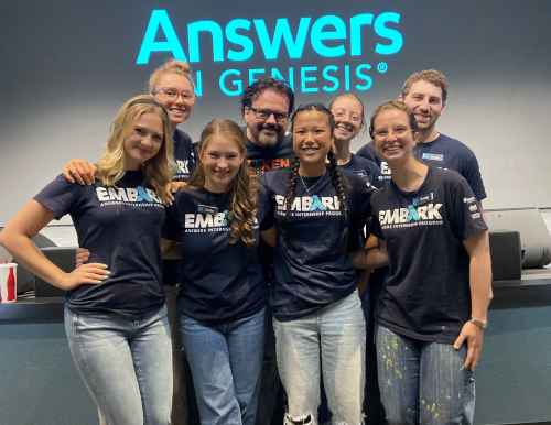 A group of eight people stands in front of a wall with the words “Answers in Genesis.” Most wear matching “Embark” shirts, smiling at the camera as they gather to celebrate the glory of God. The group includes both men and women.