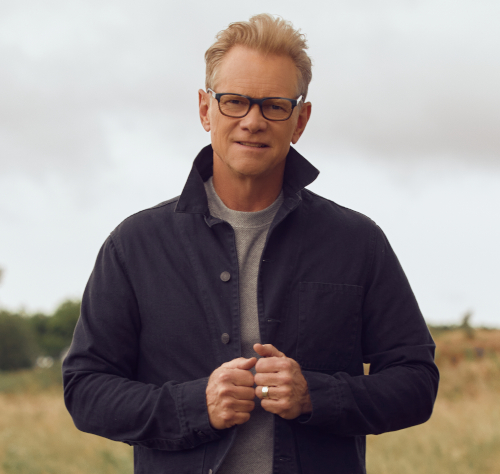 A middle-aged man with short blond hair and glasses stands outdoors at a Scholarship Banquet, wearing a dark jacket over a gray shirt. He looks at the camera, holding his jacket’s edges, with the grassy background slightly blurred.