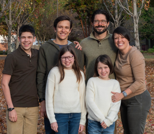 A group of six people, including two adults and four children, stand close together outdoors on a leaf-covered ground with trees in the background, all smiling at the camera—among them is Shatzer, provost at Union University.