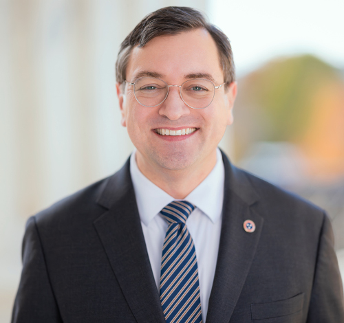 A man with short dark hair and glasses smiles while wearing a dark suit, striped tie, and white shirt. He stands outdoors with a blurred background, ready to deliver a Union lecture on Constitution Day.