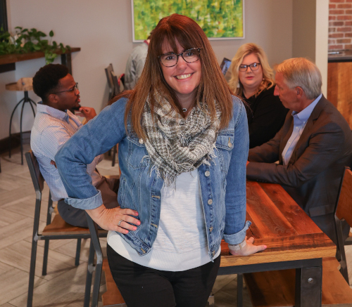 A woman in a denim jacket and scarf stands smiling by a wooden table at Union University. Behind her, two men and a woman sit engaged in conversation in this casual indoor meeting space, perfect for adult degree programs.