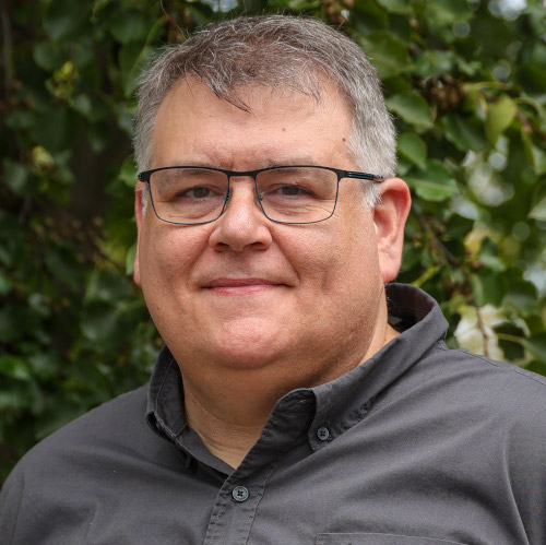Stanfield, dean of the Union School of Social Work, is a middle-aged man with short gray hair and glasses, wearing a dark button-up shirt and standing outdoors in front of green foliage. He faces the camera, smiling slightly.