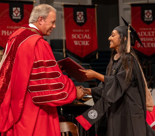 A university official in a red academic gown shakes hands with a smiling summer graduate in a black cap and gown while handing her a diploma. Christ-centered banners for different colleges hang in the background.