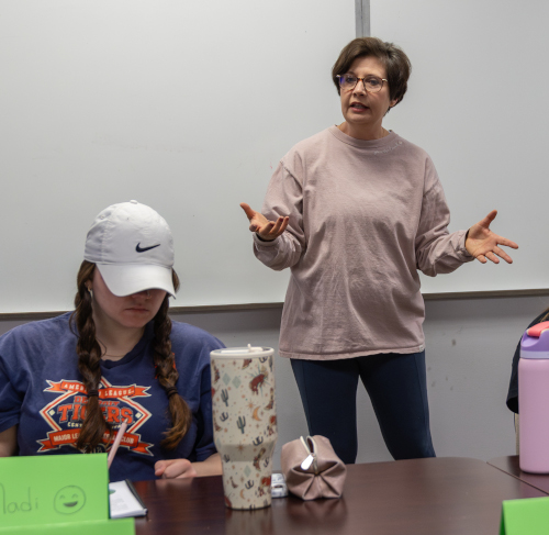 A woman stands and speaks with her hands raised in front of a whiteboard, leading a mentoring program while another person wearing a white cap sits at a table with large drink tumblers and research papers in front of her.