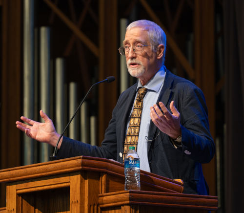 An older man with gray hair and a beard speaks at a wooden podium, gesturing as he discusses early American divisions. A bottle of water rests nearby, and large organ pipes stand in the background, emphasizing his call for national unity.