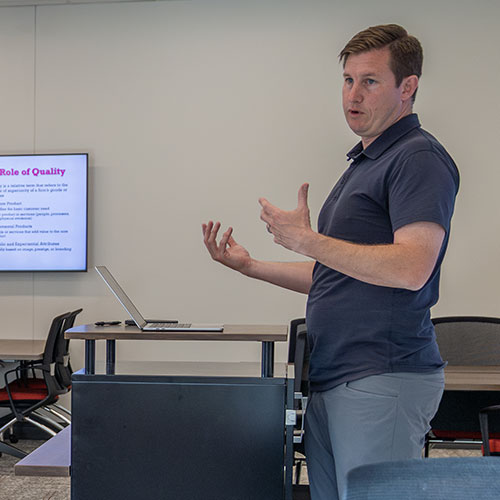 A man stands at a podium giving a presentation in an office or classroom. He gestures with his hands while speaking. A laptop is open on the podium, and a screen behind him displays a slide titled Role of Quality at a Christian university.