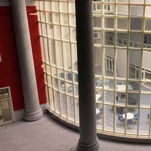 A large curved glass window with a grid pattern overlooks the New Academic Building interior with tables and chairs. Two gray columns stand in front of the window, and a red brick wall is on the left side.