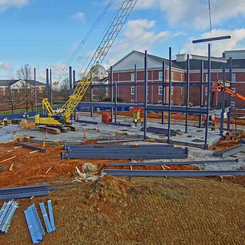 A construction site with steel beams assembled into a building framework, a yellow crane lifting a beam, workers in safety gear, and heavy equipment—one of the Top Photos 2023 set against brick buildings under a partly cloudy sky.