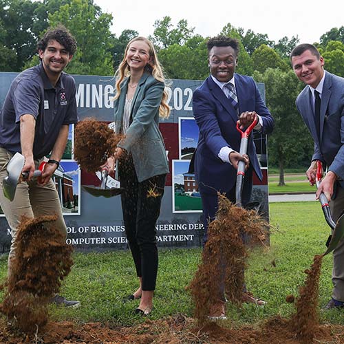 Four people in business attire use shovels to turn over dirt at a groundbreaking ceremony outdoors, featured among the Top Photos 2022, with a large sign and green trees visible in the background.
