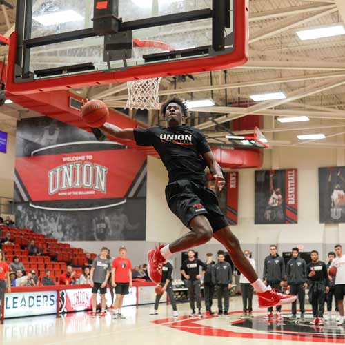 A basketball player in a black Union shirt leaps for a dunk in an indoor gym. Other players and coaches watch nearby, with banners and a red-and-black Union sign—a moment featured among the Top Photos 2021—in the background.