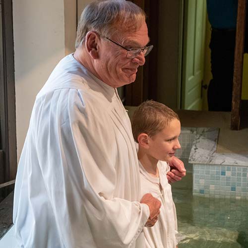 An older man and a young boy, both dressed in white robes, stand beside an indoor baptismal pool, preparing for a baptism ceremony—a touching Union story of giving back across generations.