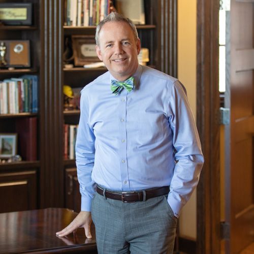 A man wearing a light blue dress shirt, green and blue bow tie, and gray pants stands in an office with wooden bookshelves and a desk, smiling at the camera—exuding leadership fitting for a college president during the COVID-19 pandemic.