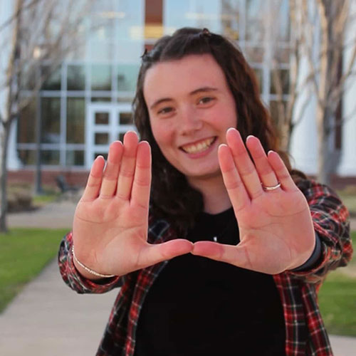 A person with long curly hair smiles while holding up both hands in a “U” shape, offering student advice. They are standing outside in front of a building with large windows.