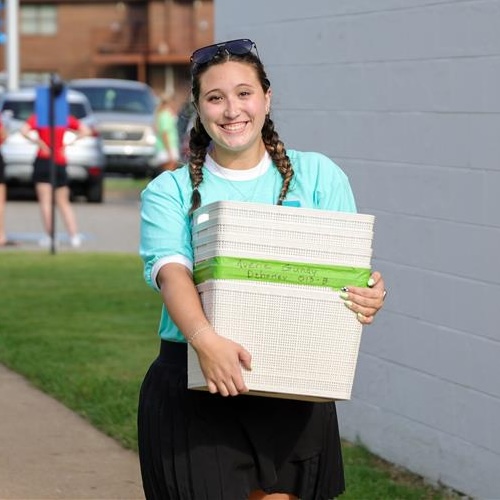 A young woman with braided hair, wearing a turquoise shirt and black skirt, smiles while carrying a stack of beige storage bins outdoors—perfect for college parenting tips near a white wall and parked cars.