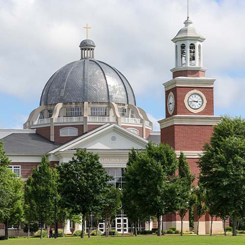 A large brick building with a domed roof topped by a cross and a clock tower, partially obscured by green trees under a cloudy sky—one of the Top Photos 2024.
