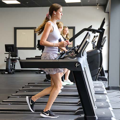Three women are exercising on treadmills in a gym, embracing new health tips to be healthier in 2025. The woman in the foreground wears a tank top, shorts, and sneakers, with wireless earbuds and a scrunchie. Other gym equipment appears in the background.