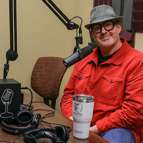 A man wearing a gray fedora, black glasses, and a red jacket sits at a podcast recording table, ready to share tips on starting a podcast. A microphone, headphones, tumbler, and podcast equipment are visible.