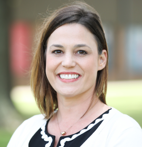 A woman with straight brown hair and a white top with black trim stands outdoors, smiling at the camera—recently appointed dean at Union College of Education. The background is blurred with some trees and indistinct structures.