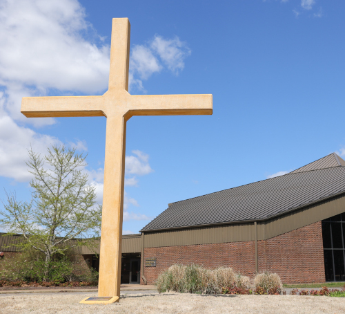 A large yellow cross stands in front of a brick building with a brown roof, where Union students often develop their preaching skills, surrounded by sparse landscaping and under a partly cloudy blue sky.