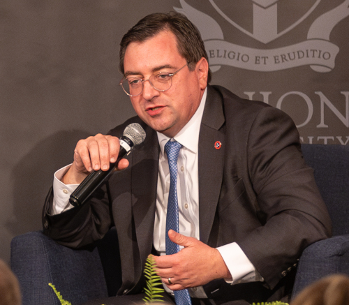 A man in a suit and tie is sitting in a chair, holding a microphone and speaking about law. He wears glasses and has short brown hair. The dark background features a partially visible emblem and text.