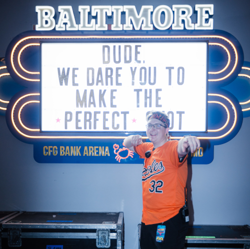 A Union student in an Orioles jersey points toward the camera while standing in front of a lit sign that says, “Dude, we dare you to make the perfect shot” at CFG Bank Arena in Baltimore.