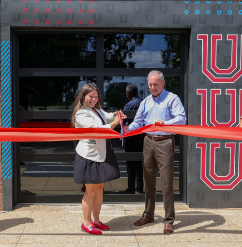 Two people stand outside a building with stylized letter U designs, holding oversized scissors and cutting a large red ribbon at the entrance—signaling the grand opening of the Union Wellness Center.
