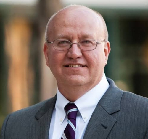 A man with glasses and a bald head is wearing a gray suit, white shirt, and striped tie. He is standing outdoors at the Union luncheon, smiling at the camera, embodying Faith in the Marketplace.