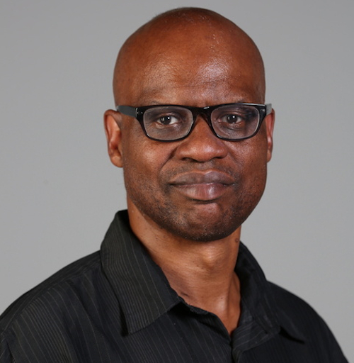 A man with a shaved head wearing black rectangular glasses and a black collared shirt stands in front of a plain gray background, looking at the camera with a neutral expression, as if ready to discuss topics like racism or participate in the Jones Lecture.
