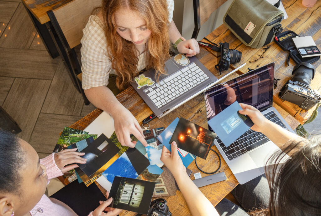 Three people sit at a wooden table sorting through printed photos, embodying the collaborative spirit of photojournalism. A laptop, cameras, a lens, and a green bag—all essential tools in digital media communications—are scattered across the table. The scene is lit by natural light from the left.