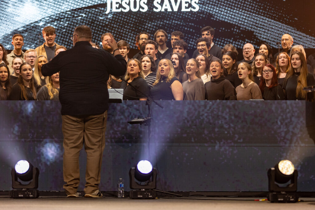 A choir of men and women sings on stage, facing a conductor, exemplifying the power of music education. The words JESUS SAVES are displayed above them. Stage lights and a microphone are visible in the foreground.