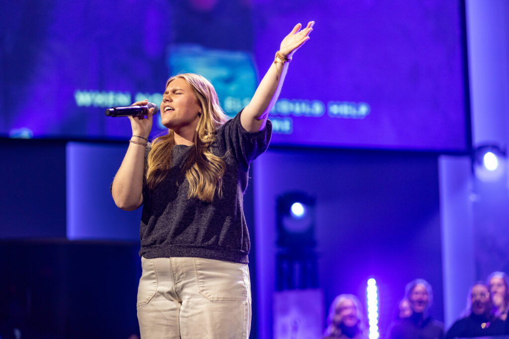 A woman with long blonde hair gives a powerful vocal performance, singing into a microphone on stage with one arm raised. She is wearing a dark top and light pants, while several people and purple lighting highlight this music education event.