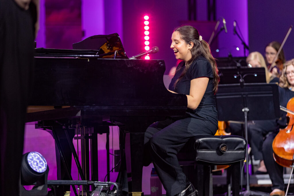 A woman dressed in black sits at a grand piano, singing into a microphone during a captivating music performance. Behind her, several musicians play string instruments on a stage illuminated by purple lights.