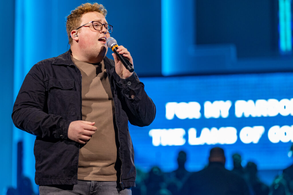 A person with glasses sings into a microphone on stage during a music performance. They are wearing a dark jacket and beige shirt. Blue lighting and an out-of-focus screen with white text appear in the background.