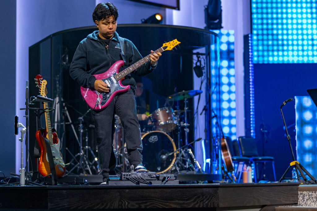 A person in a dark hoodie and jeans plays an electric guitar on stage, showcasing live performance technology. A drum set and other guitars are visible in the background, with blue stage lights illuminating the scene.