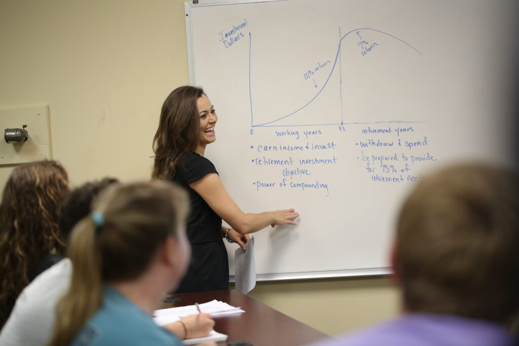 A woman stands at a whiteboard, smiling while explaining an Economics graph to a group seated at a table. The chart labeled “Investment Dollars” includes notes on retirement investment and key Business Administration concepts.