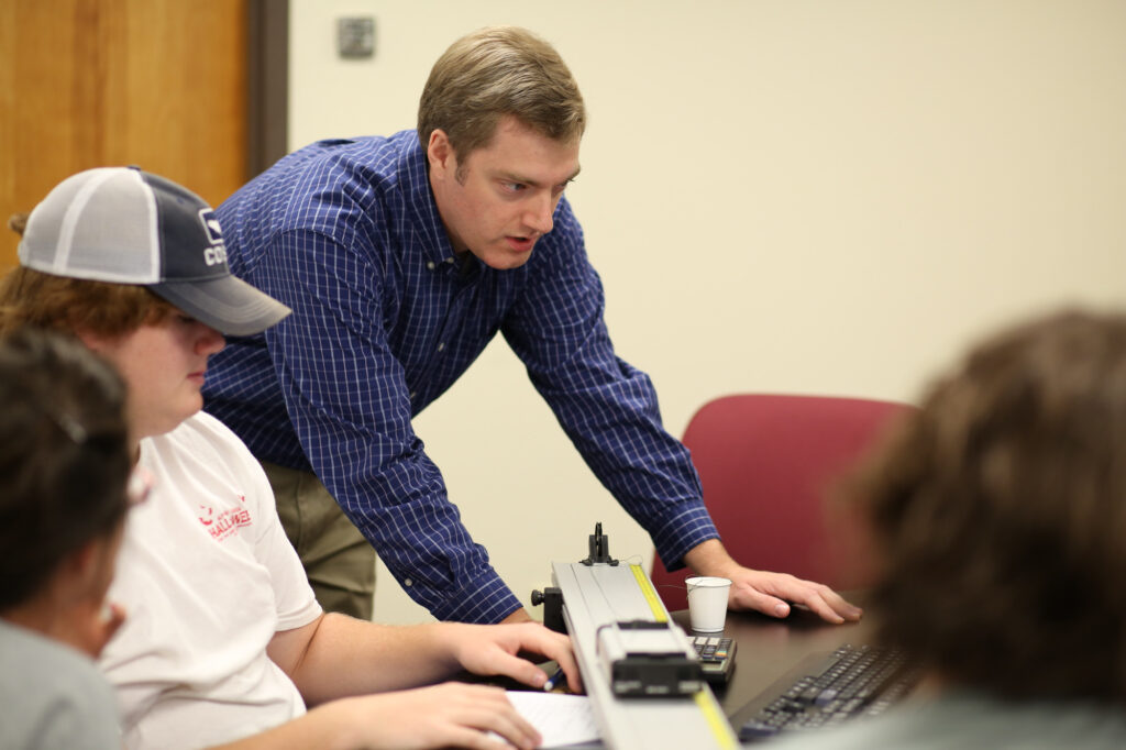 A man in a blue plaid shirt leans over a desk, instructing students who are working on a computer and using a scientific measuring device in a classroom setting.