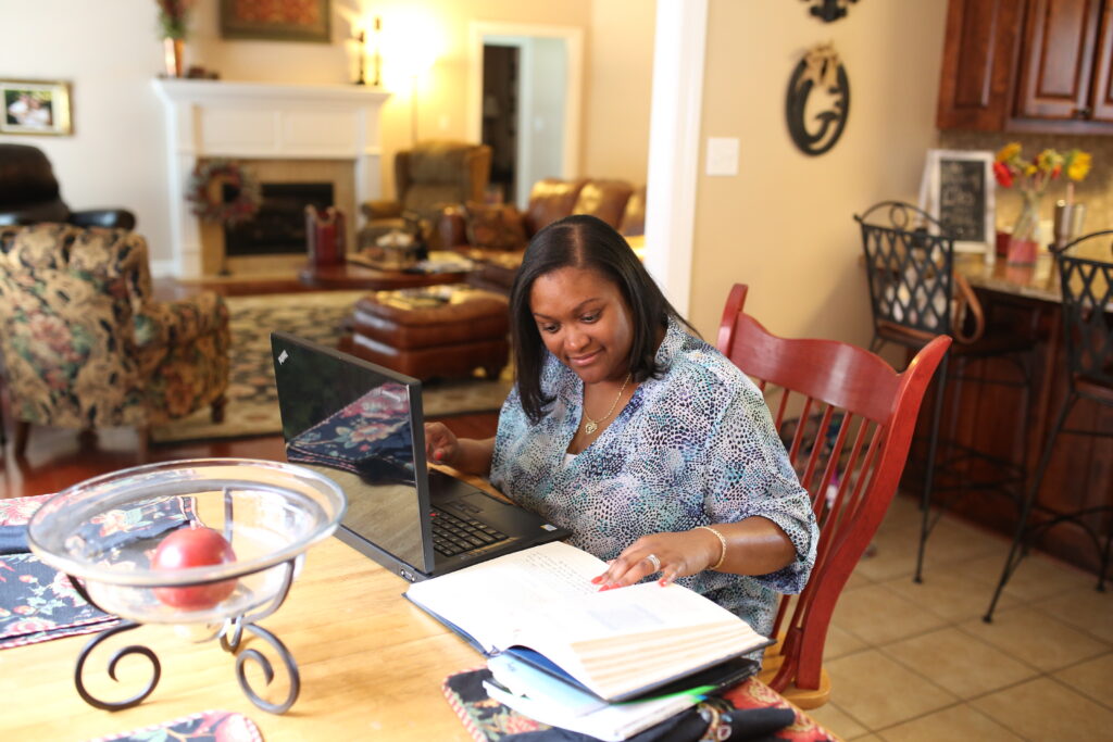 A woman sits at a wooden table in a kitchen, working on her laptop and reading an open BSOL book. A glass bowl with fruit is on the table, and the background features a living room and bar stools.