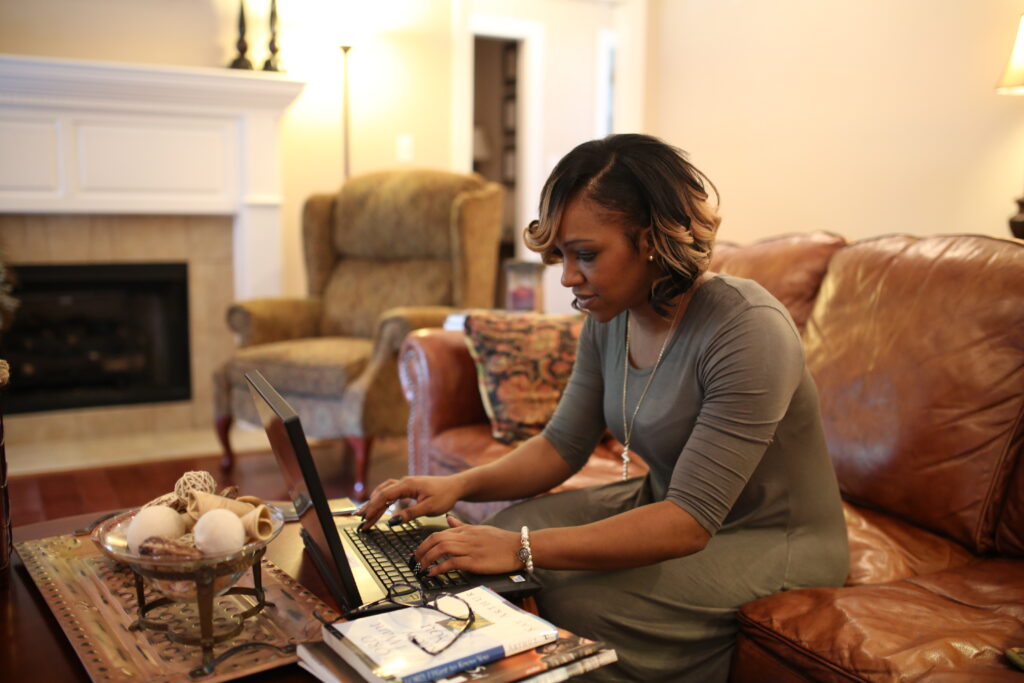 A woman sits on a brown leather sofa in a cozy living room, typing on her laptop for a BSOL Project Management task. Warm lighting, a fireplace, an armchair, and decorative items adorn the inviting space.