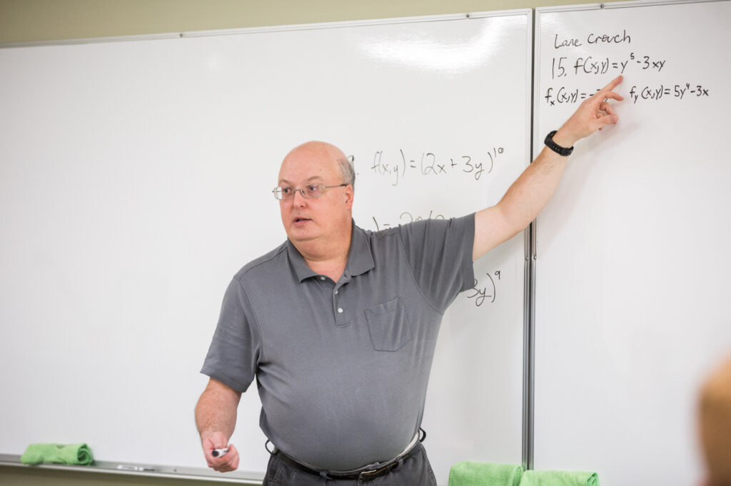 A man wearing glasses and a gray polo shirt stands in front of a whiteboard, pointing at math equations written in black marker. The board contains functions and calculus notation. Green cloths rest on the boards tray.