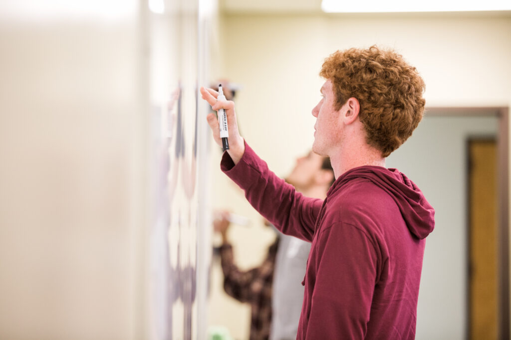 A person with curly hair wearing a maroon hoodie writes on a whiteboard with a black marker. Another person in the background is also writing on the whiteboard. The setting appears to be a classroom.